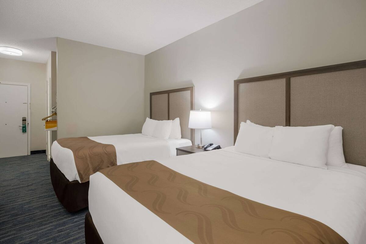 Una habitación de hotel con dos camas, una mesita de noche con lámpara y un armario para abrigos junto a la puerta.