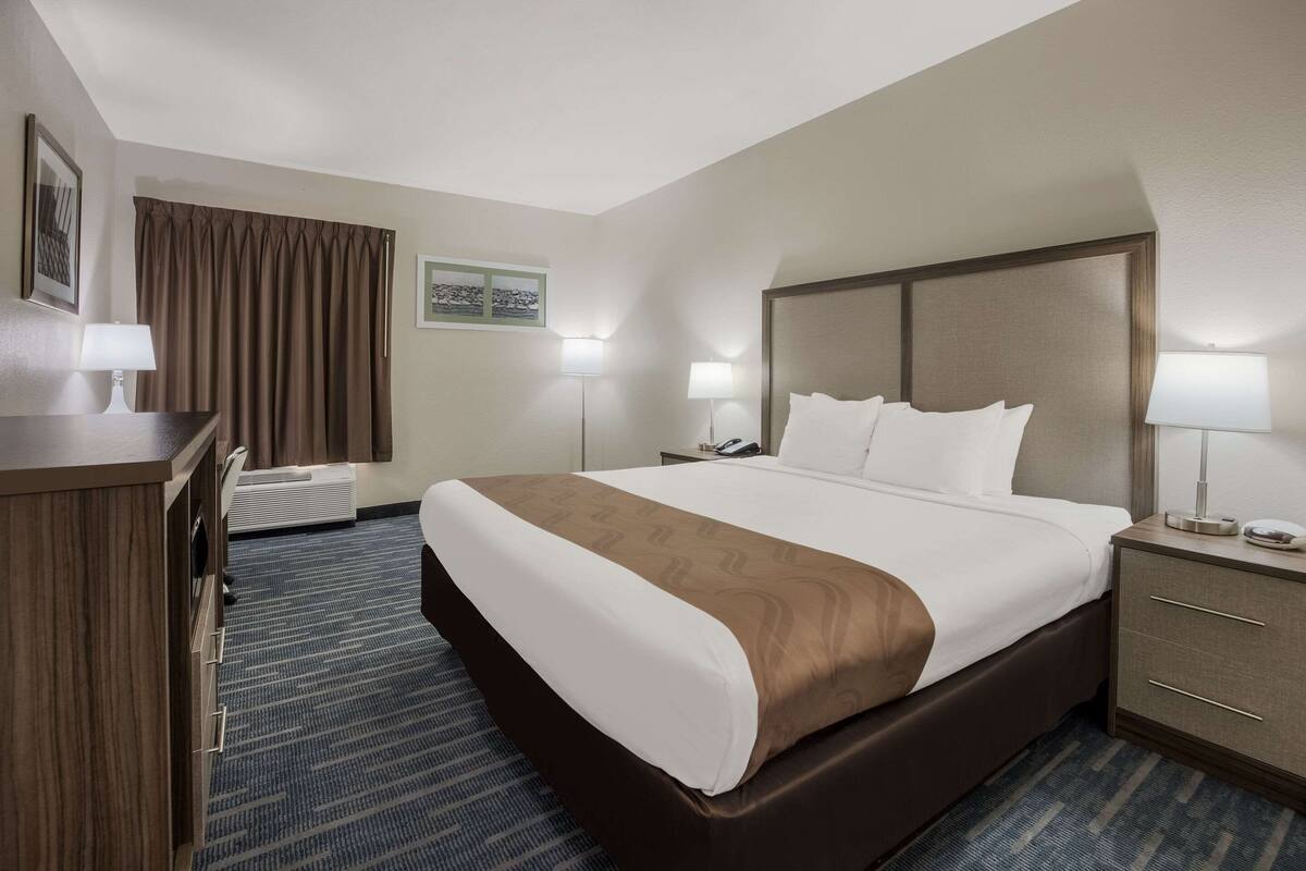 Habitación de hotel con una cama grande, dos mesitas de noche con lámparas, un escritorio con lámpara, una silla, un ventanal con cortinas y un aire acondicionado instalado en la pared.