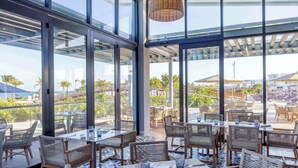 Un amplio comedor interior con ventanas grandes, mobiliario de mimbre y vistas a la playa y palmeras.