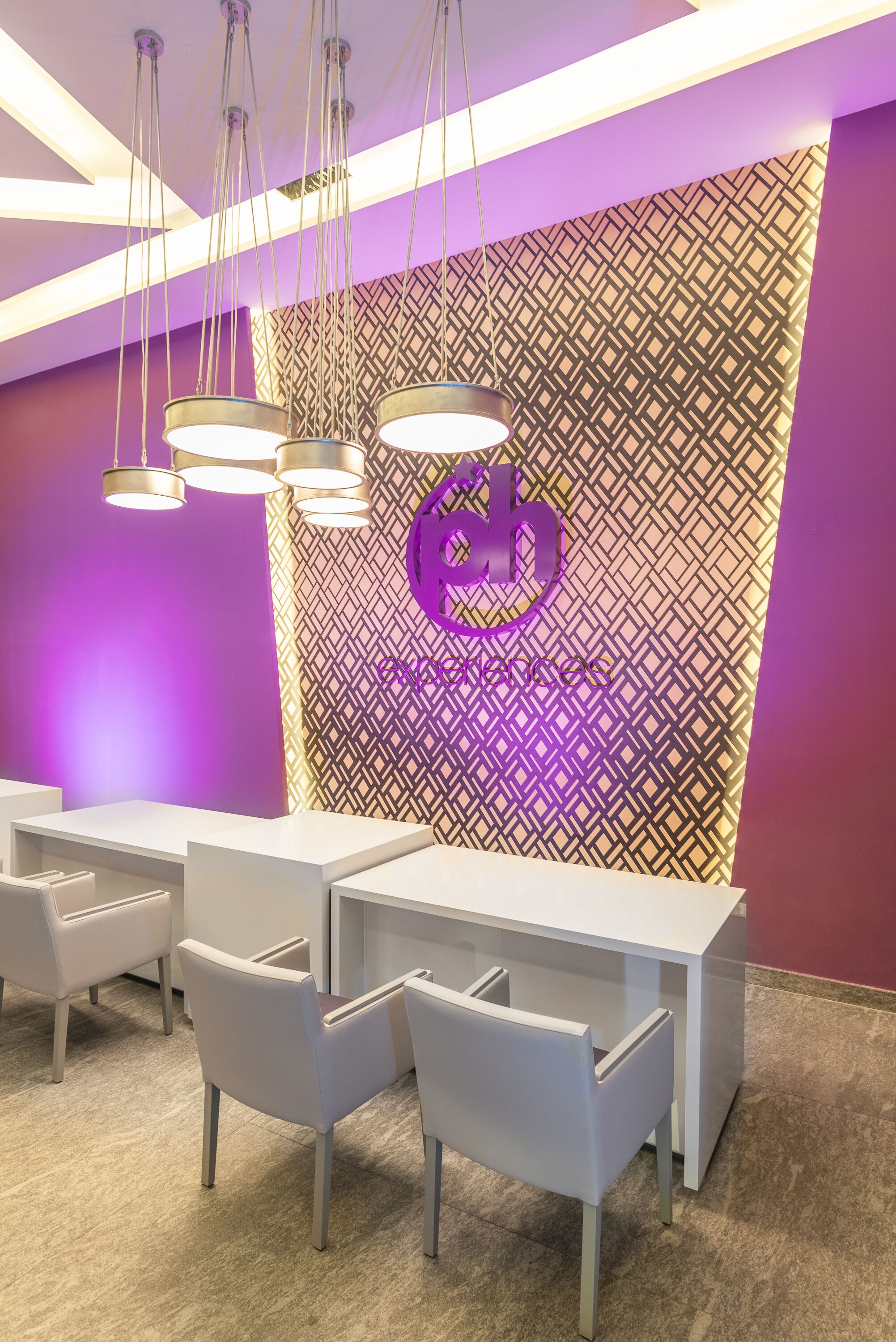 A reception area with a purple wall, a geometric patterned backdrop, and a logo. There are white tables and chairs arranged for guests.
