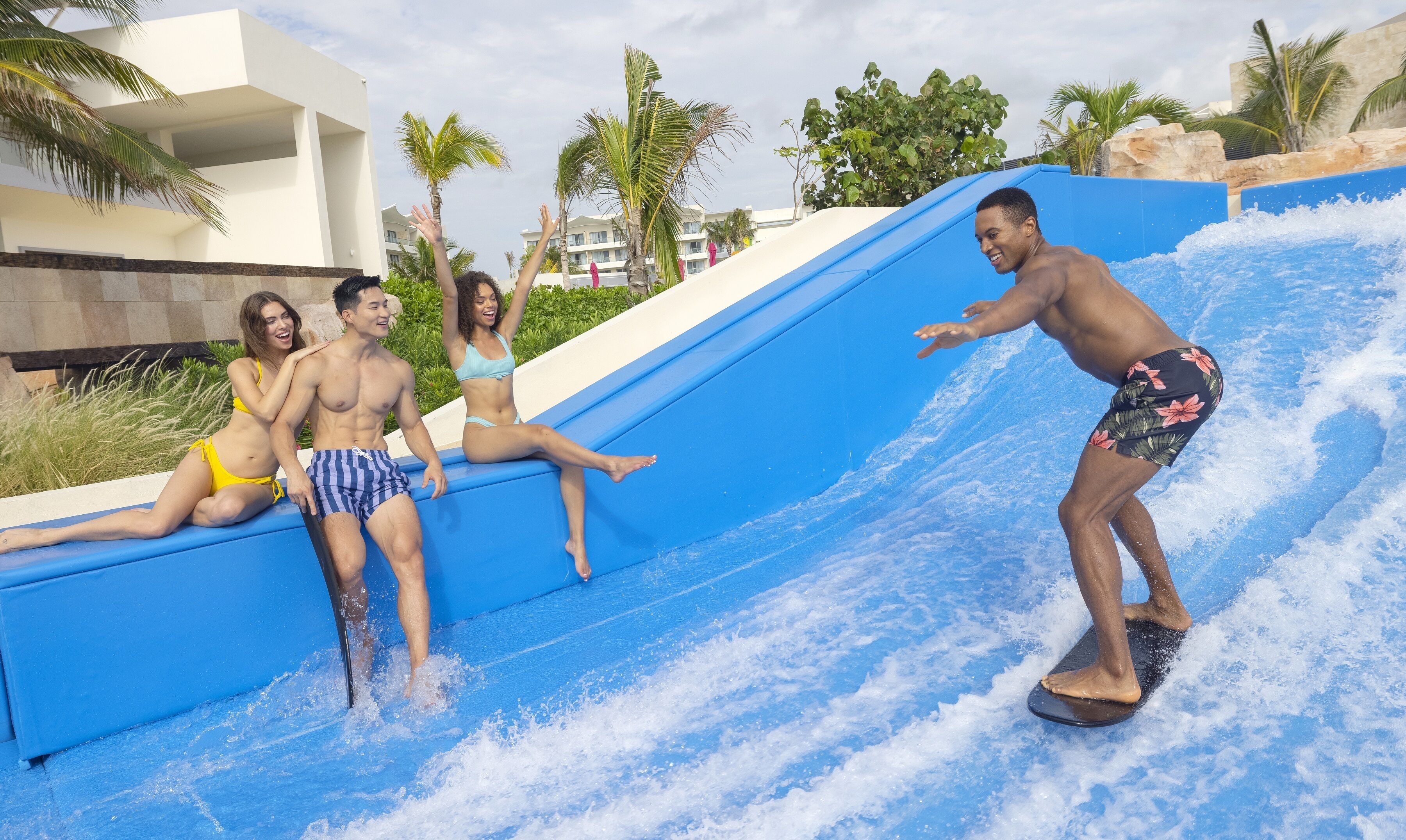 Tobogán de agua en un complejo turístico con piscina, palmeras y un edificio al fondo.