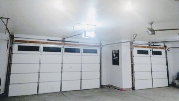 Un garage avec deux portes de garage blanches, un ventilateur de plafond et un luminaire fluorescent.