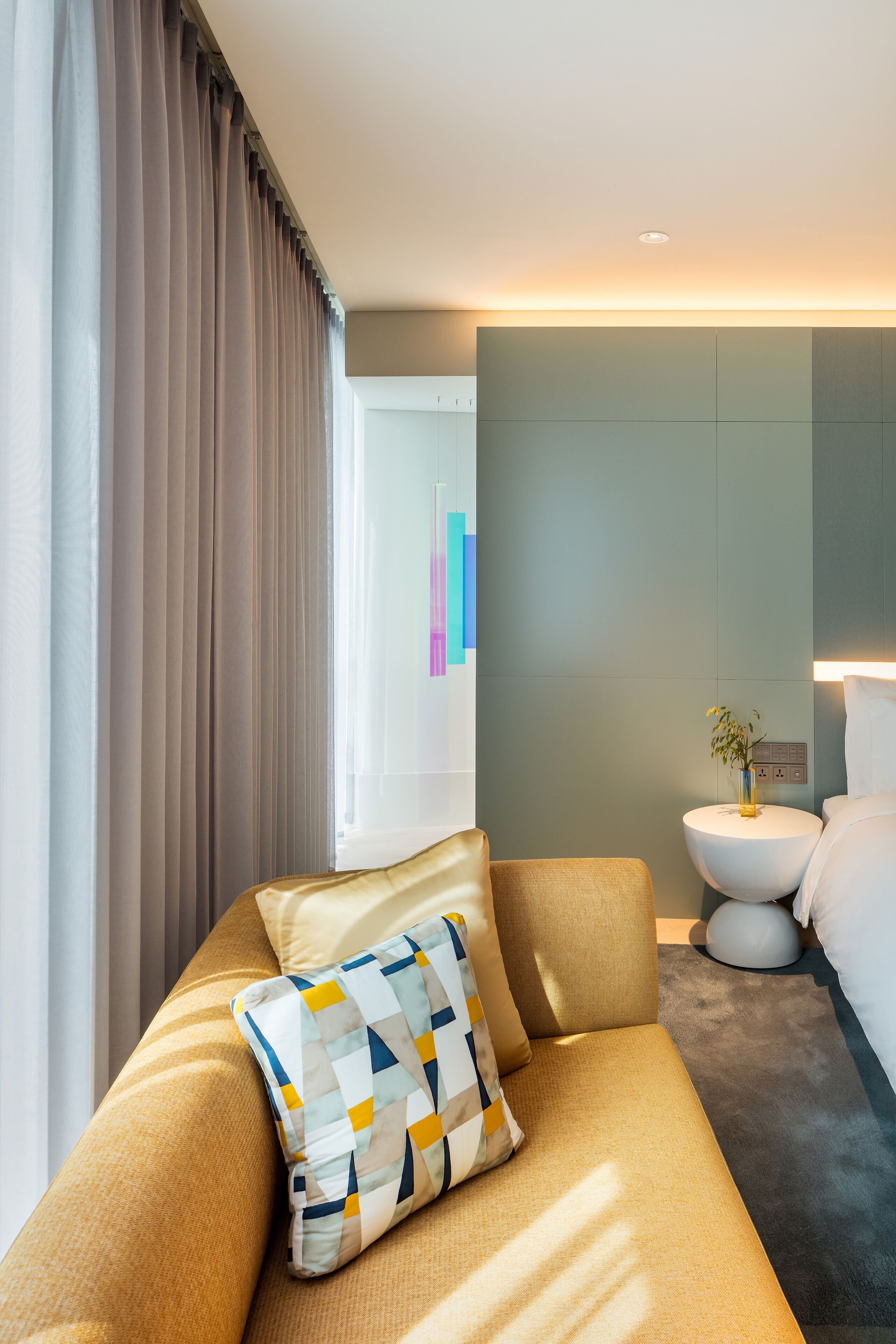 A modern hotel room with a sofa, a bedside table with a vase, and a wall-mounted light.