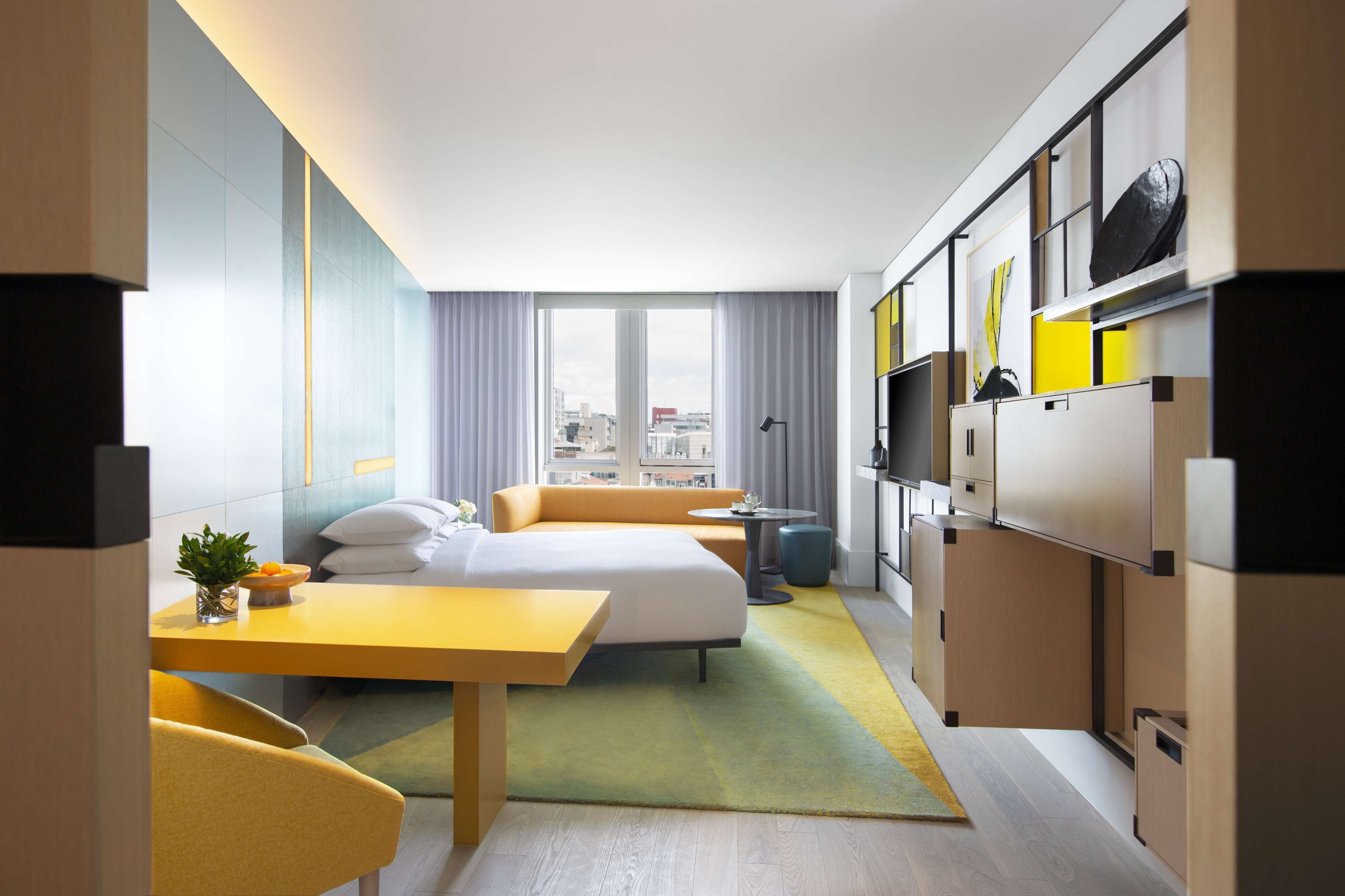 A modern hotel room with a bed, a desk, a chair, a small table, a sofa, a TV, and a wardrobe.