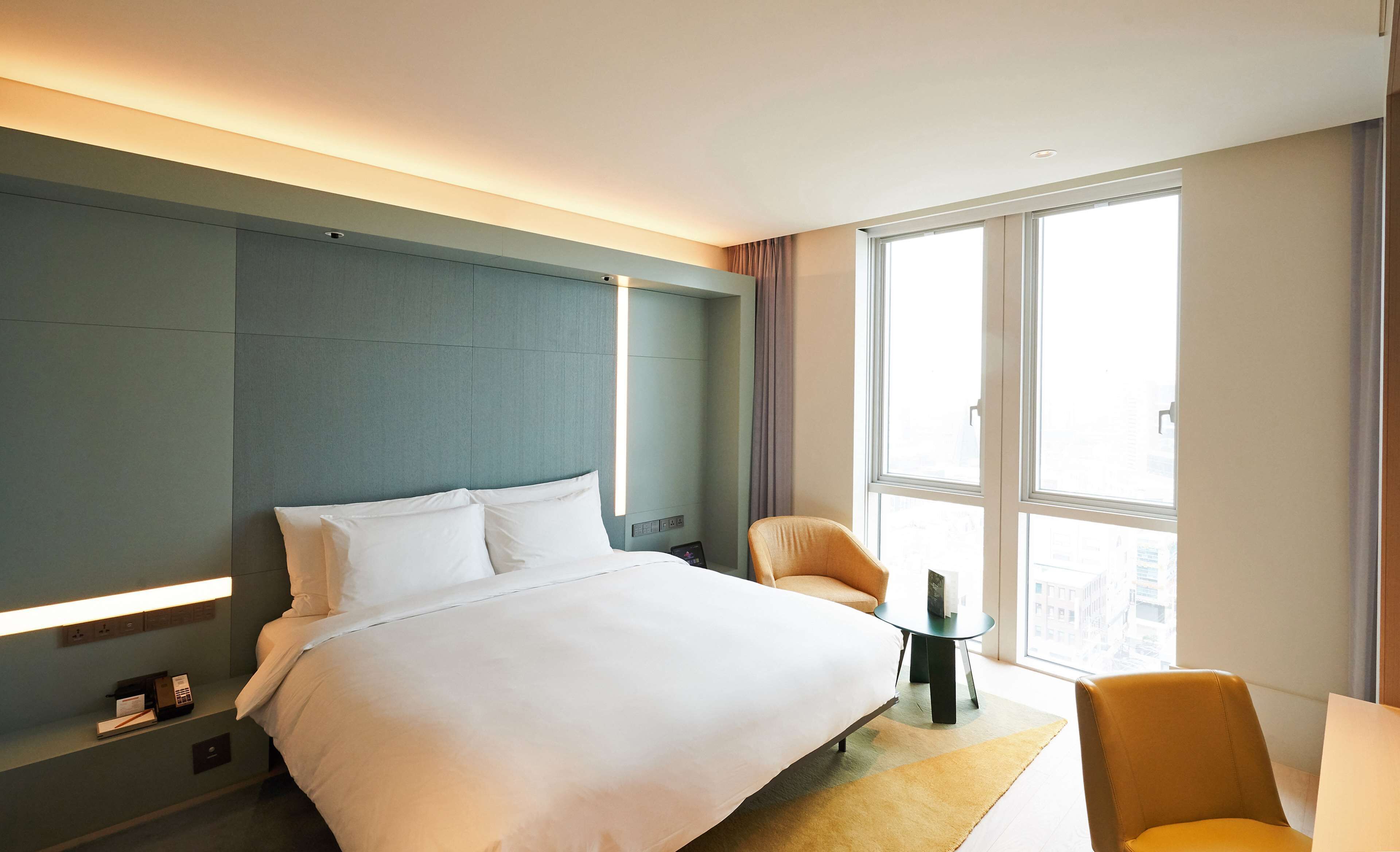 A modern hotel room with a large bed, a small table, a chair, and a view of the city through large windows.