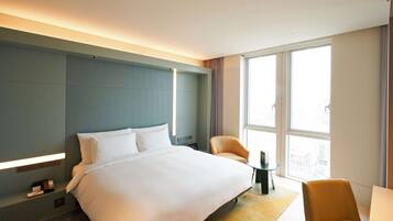 A modern hotel room with a large bed, a small table, a chair, and a view of the city through large windows.