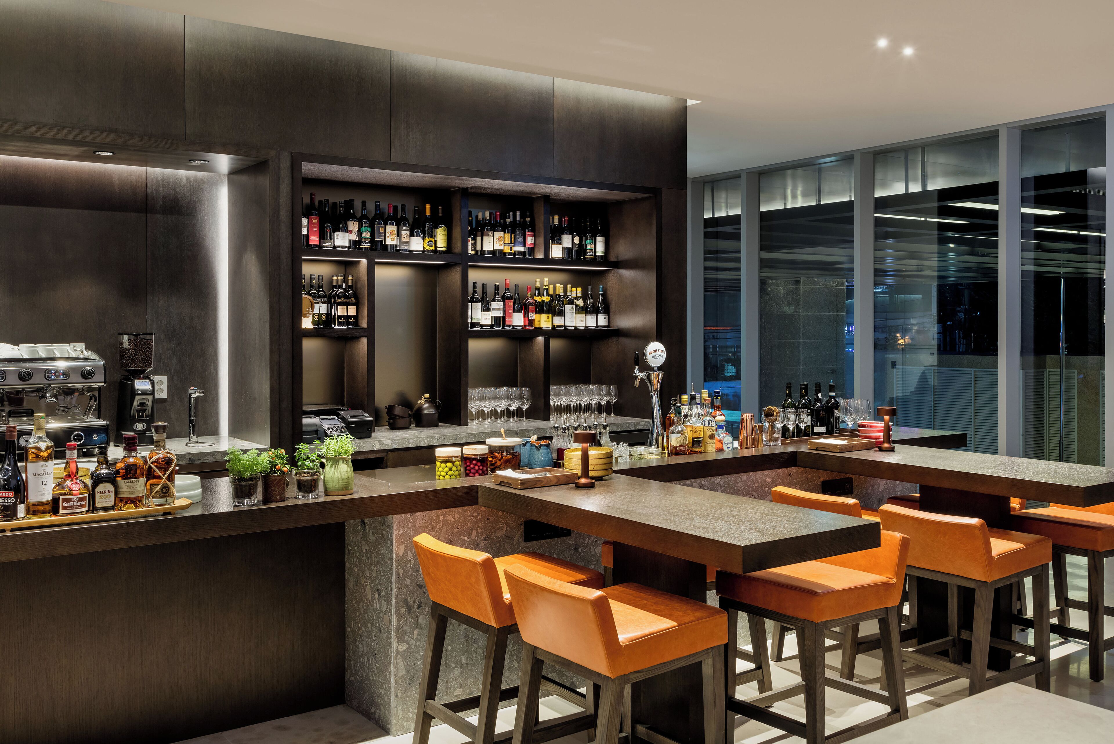 A modern bar area with a long counter, multiple shelves of liquor bottles, and several orange bar stools.