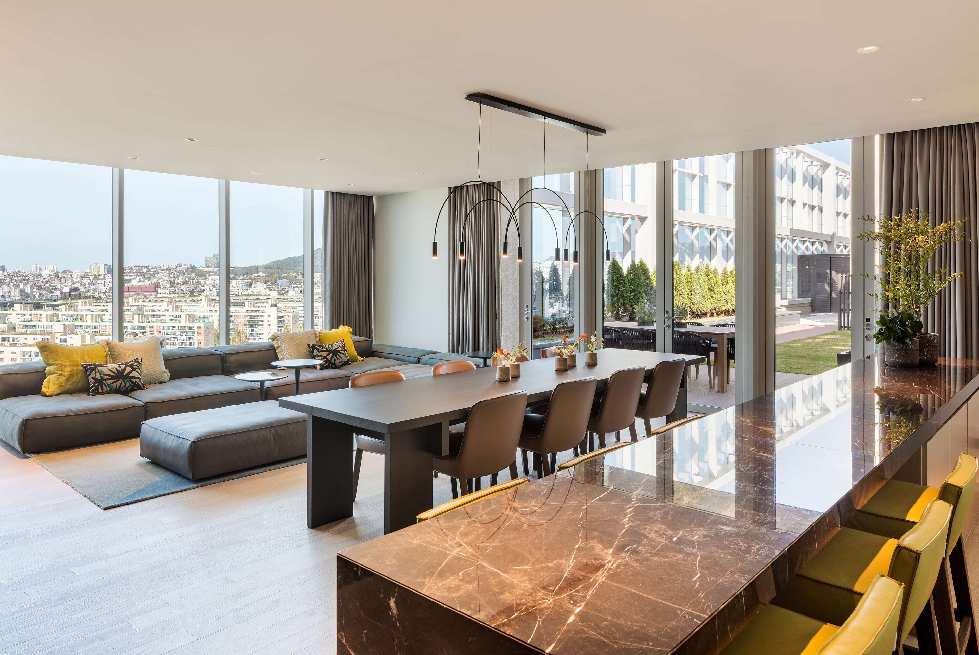 A modern living room with a large sofa, a dining table, and chairs. There is a view of a cityscape through large windows.