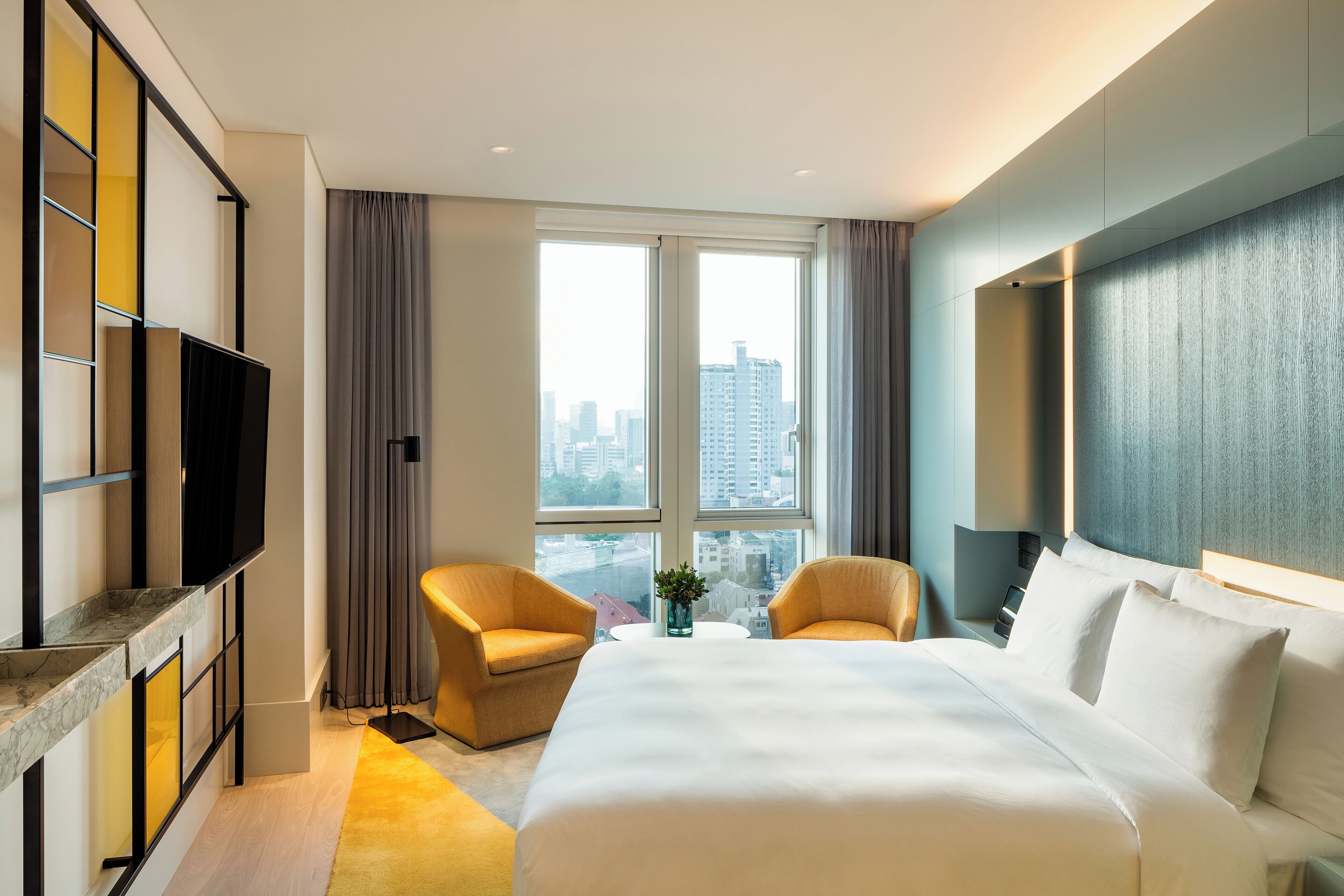 A hotel room with a large bed, two armchairs, a TV, and a city view through the windows.