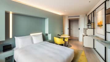 Quarto de hotel moderno com uma cama grande, uma escrivaninha com um laptop, uma mesa redonda pequena com duas cadeiras e um minibar.
