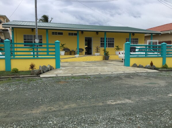 Edificio amarillo con techo verde, entrada protegida por una reja y un pequeño espacio al aire libre con mesas y sillas.