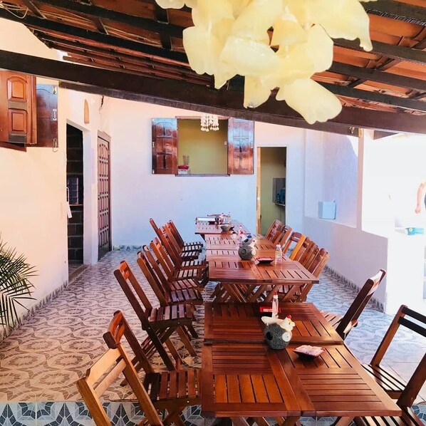 Un área de comedor al aire libre, cubierta, con mesas y sillas de madera, una gran flor decorativa y piso de baldosas.