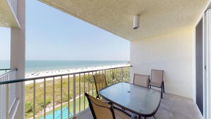 Un balcon avec une table et des chaises en verre, donnant sur une plage parsemée de parasols et fréquentée par des gens.