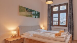 Makuuhuoneessa on sänky, yöpöydät, taulu, ikkuna verhoineen ja näkymä ulkona oleviin rakennuksiin.