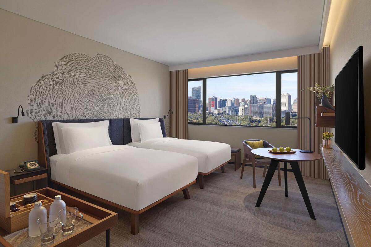 A hotel room with a bed, a desk, a chair, a small table with a bowl of fruit, a TV, and a large window with a city view.