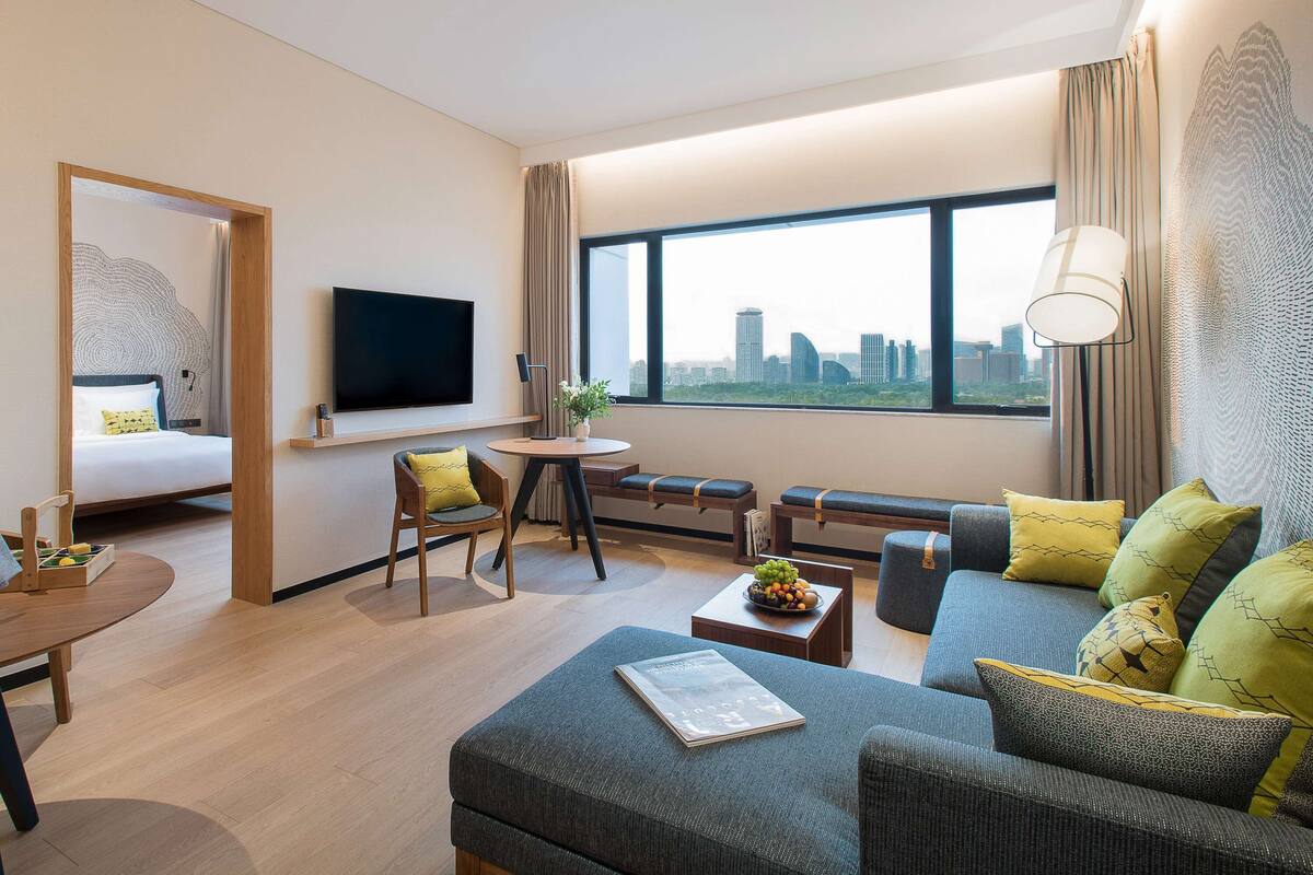 A modern hotel room with a bed, a sofa, a dining area, and a large window with a city view.