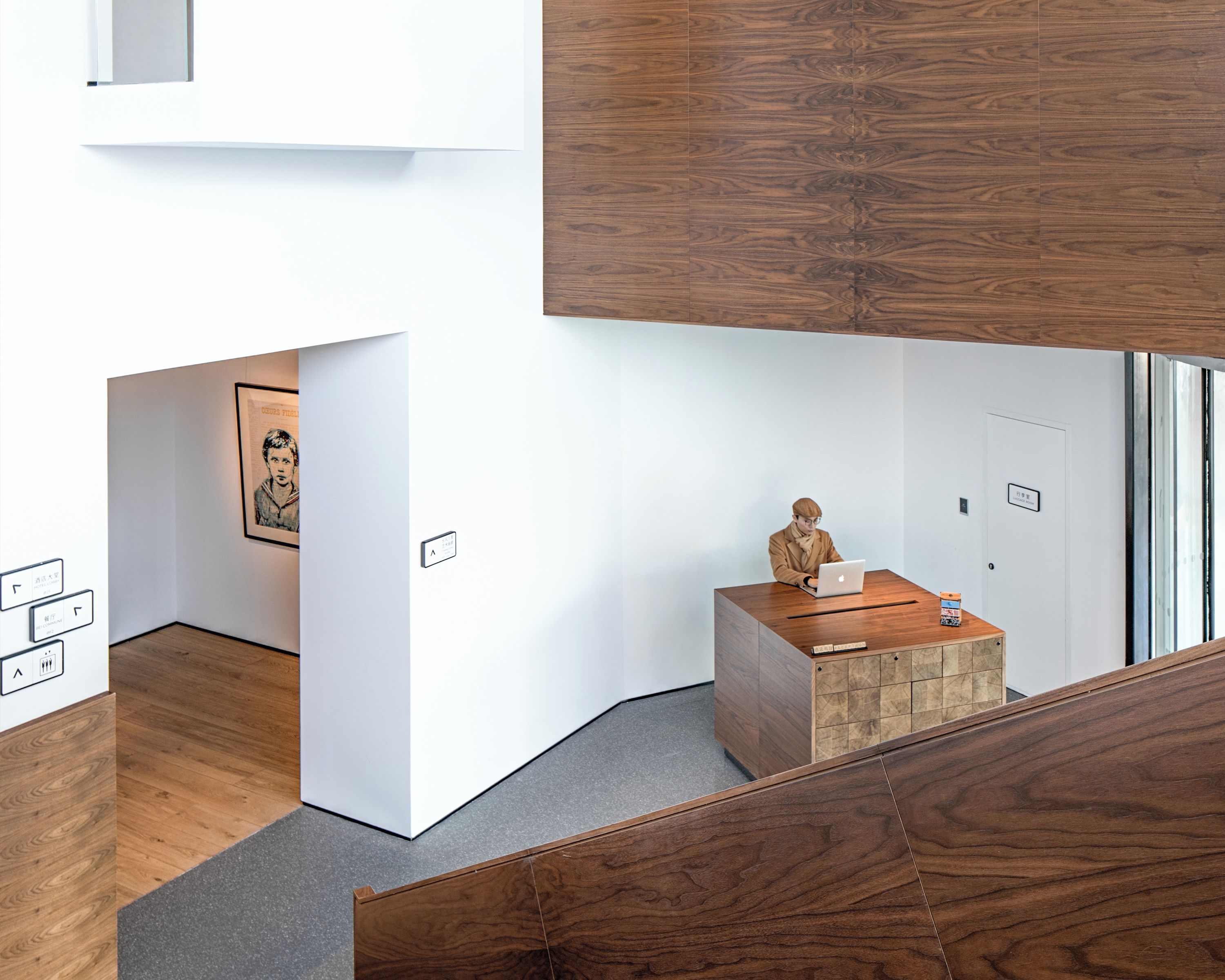 A modern hotel lobby with a wooden reception desk, a staircase, and a framed artwork on the wall.