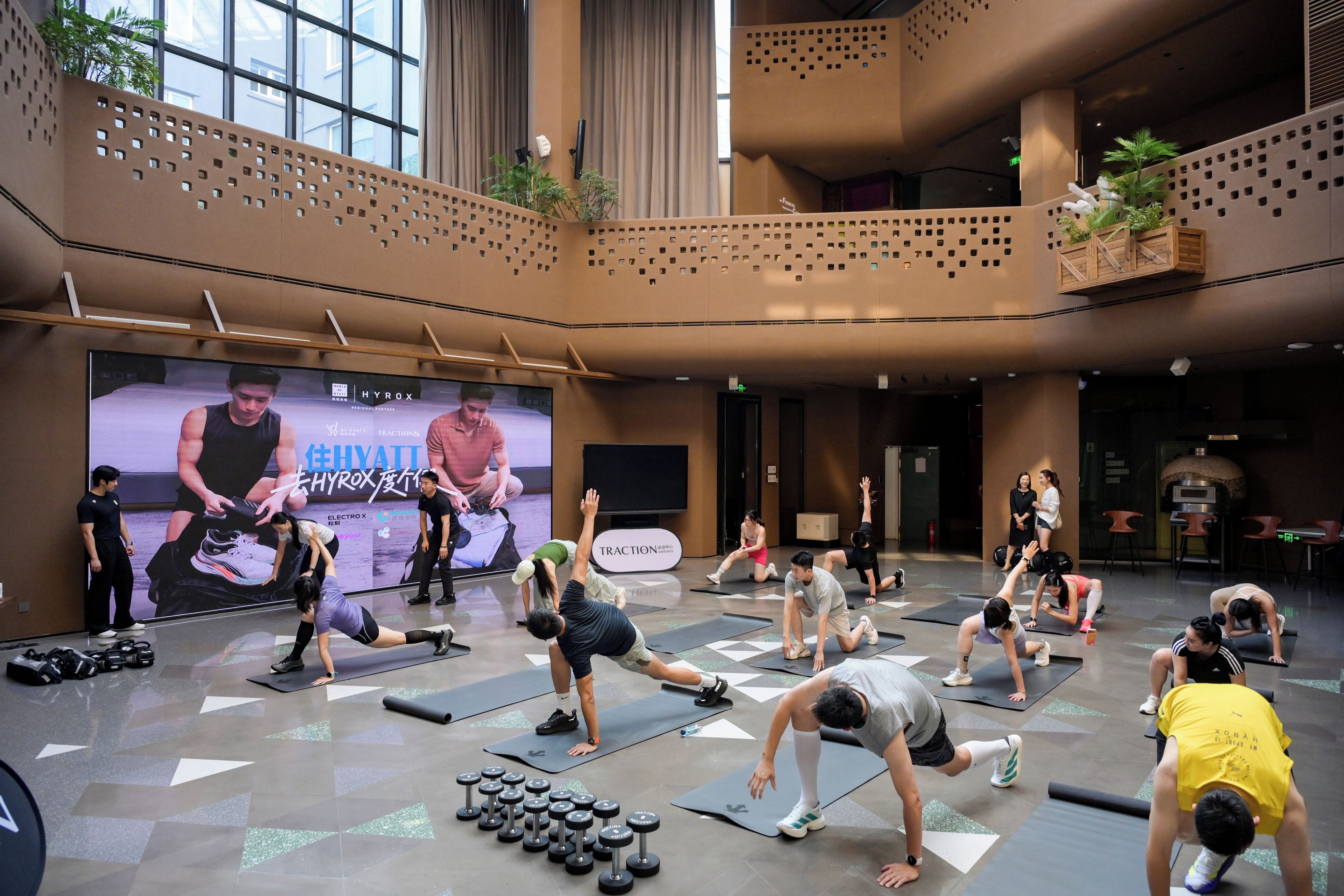 A spacious indoor gym with multiple exercise mats, dumbbells, and a large screen displaying a fitness event.