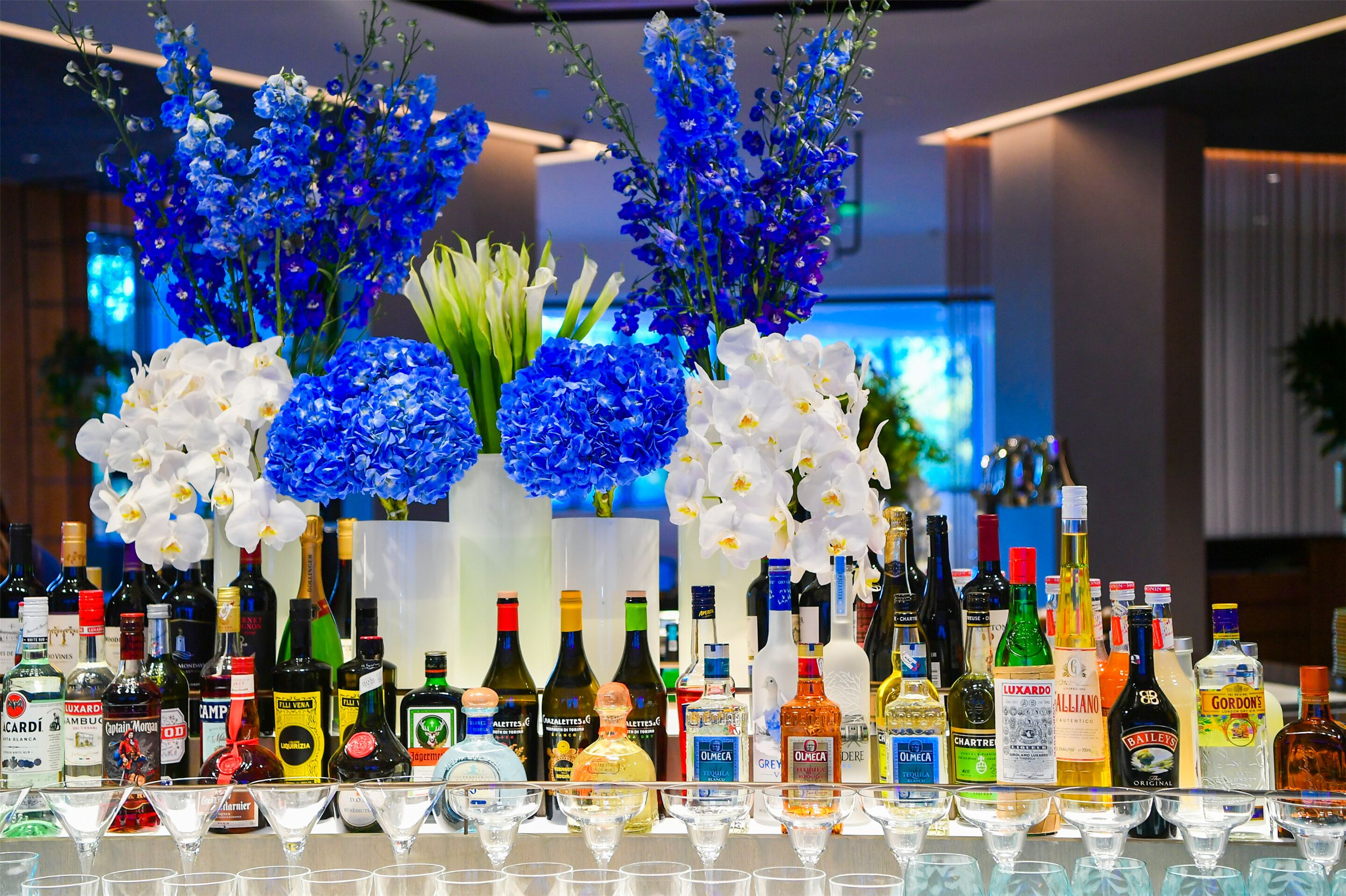 Zona de bar con diferentes botellas de licor y vasos, y un arreglo floral con flores azules y blancas.
