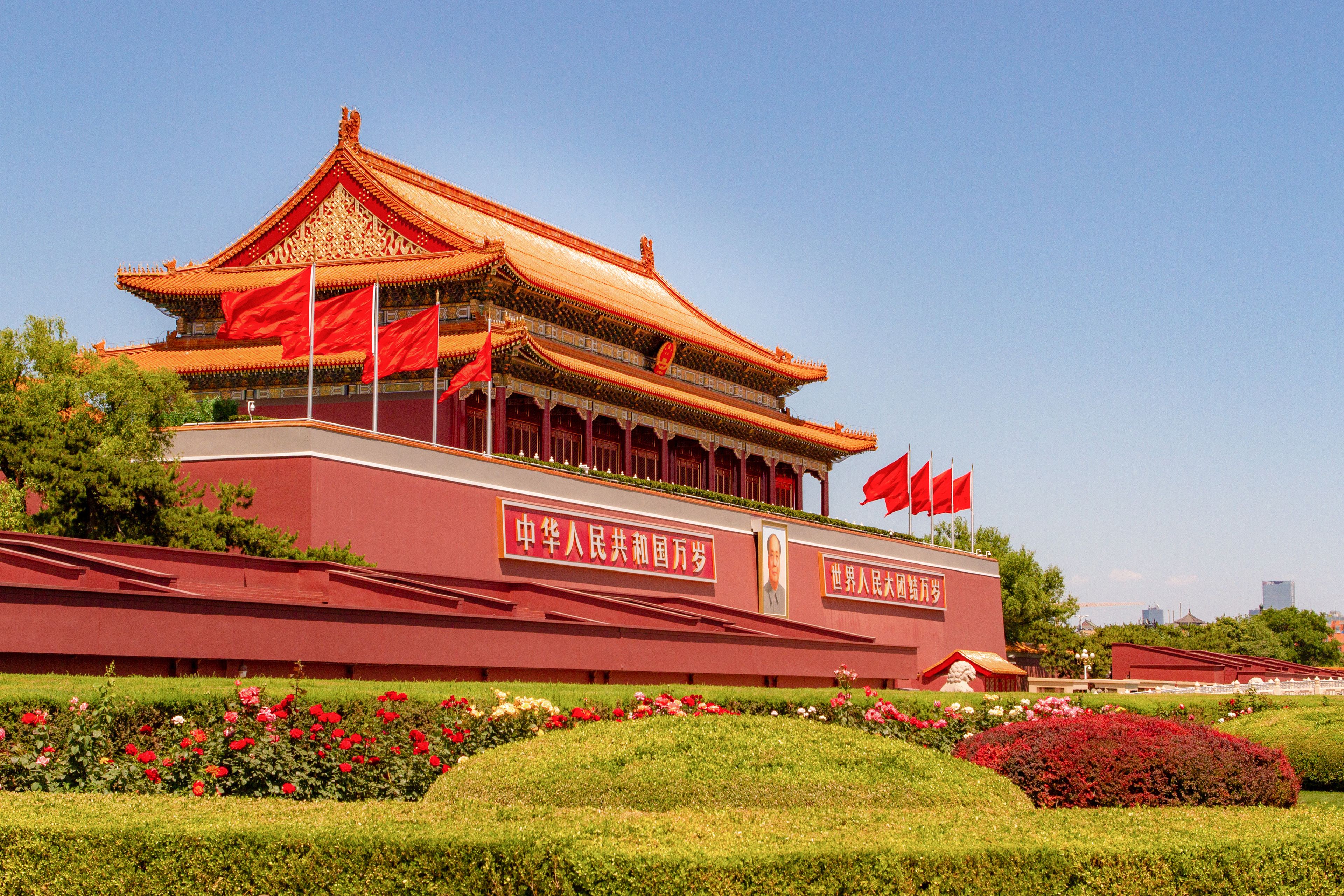 A traditional Chinese building with multiple flags, surrounded by well-maintained gardens and a clear sky.