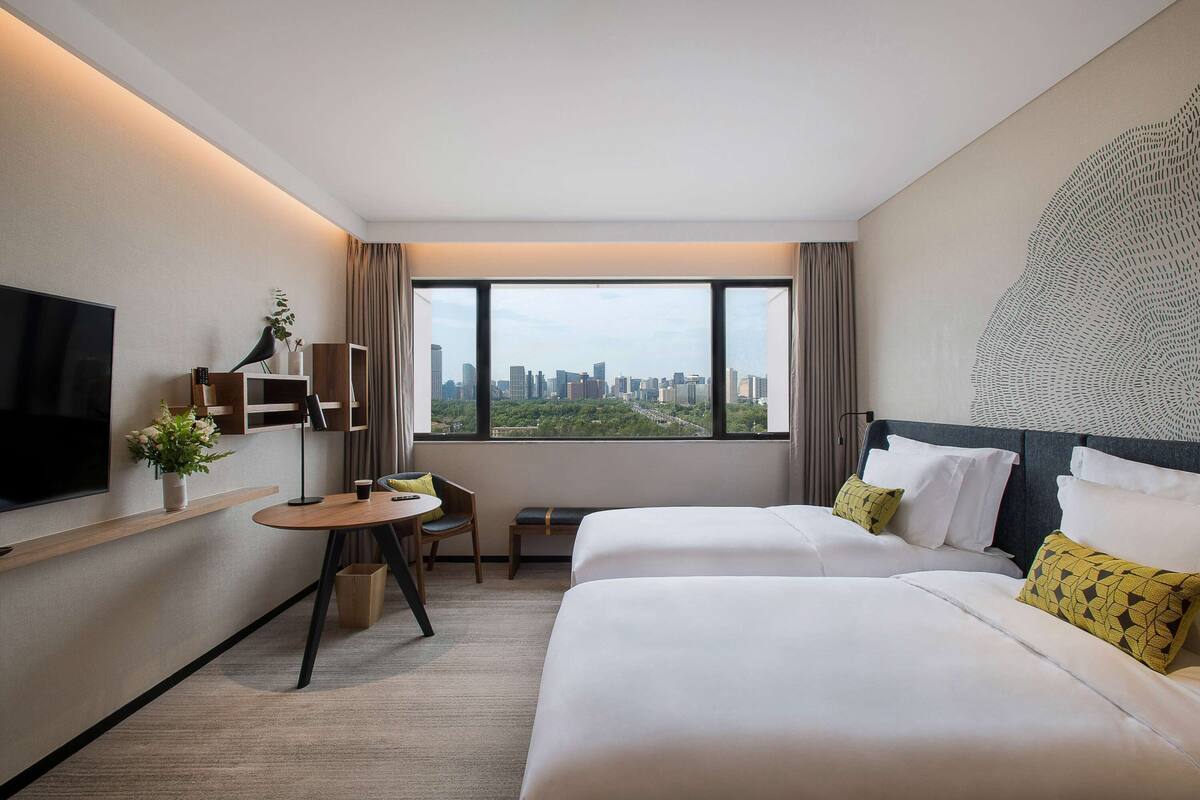 A hotel room with two beds, a TV, a small table with a chair, a window with a city view, and a decorative wall piece.