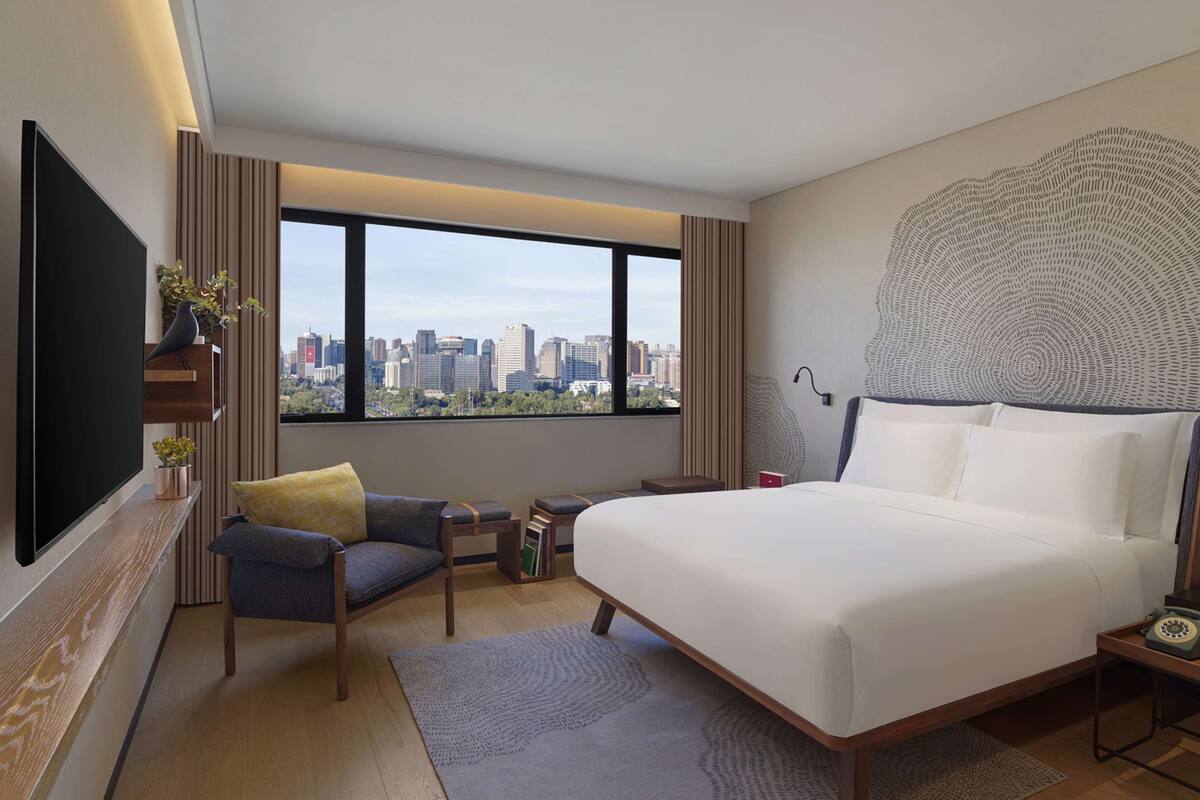 A modern hotel room with a large bed, a TV, a city view, and a unique wall art piece.