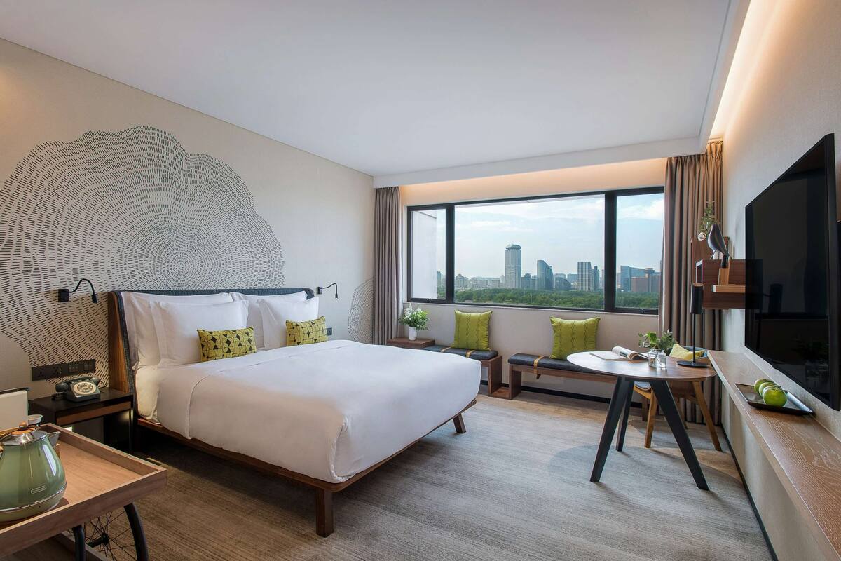A modern hotel room with a large bed, a desk, and a view of the city skyline.