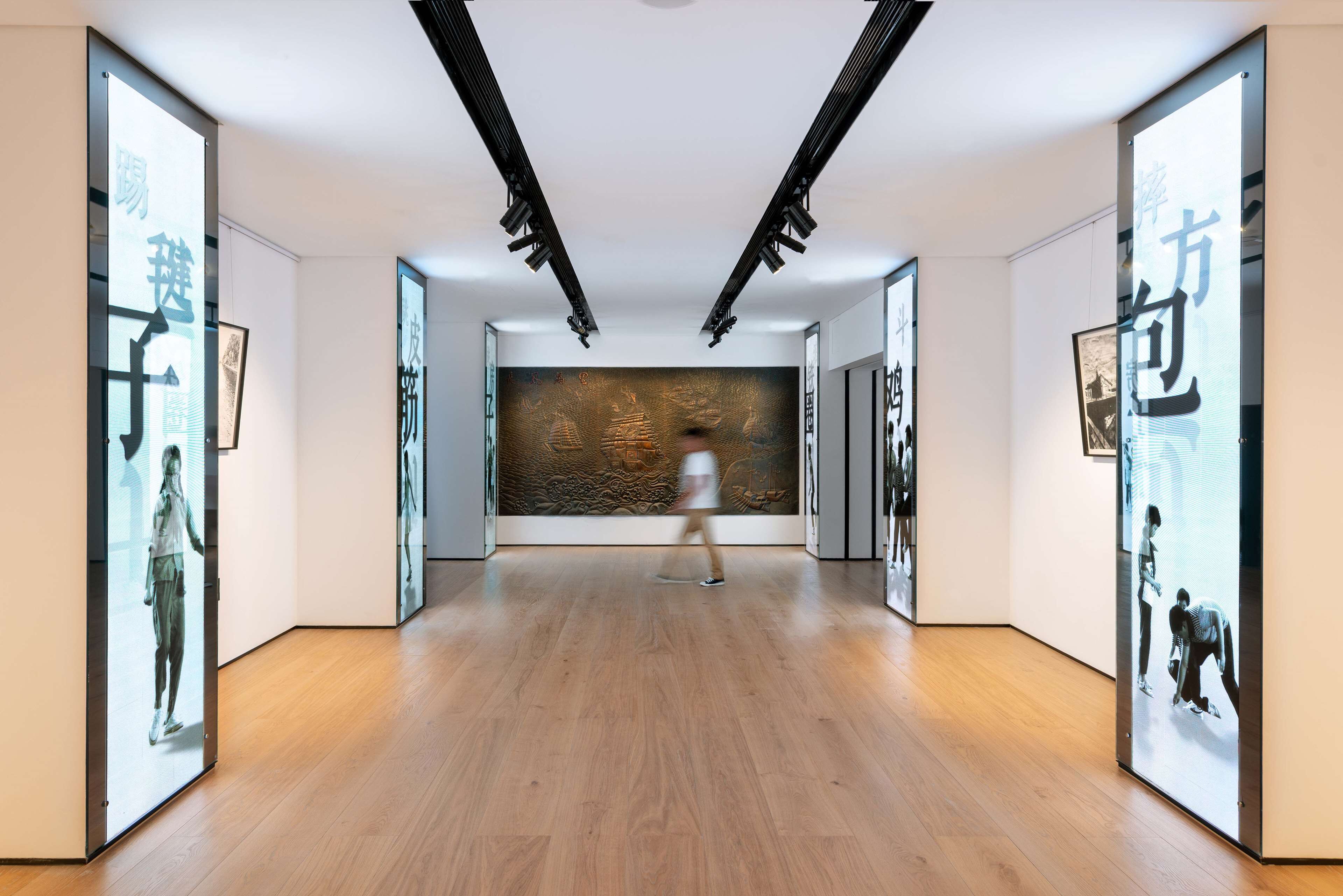 A hallway with wooden flooring, framed artwork on the walls, and a person walking.