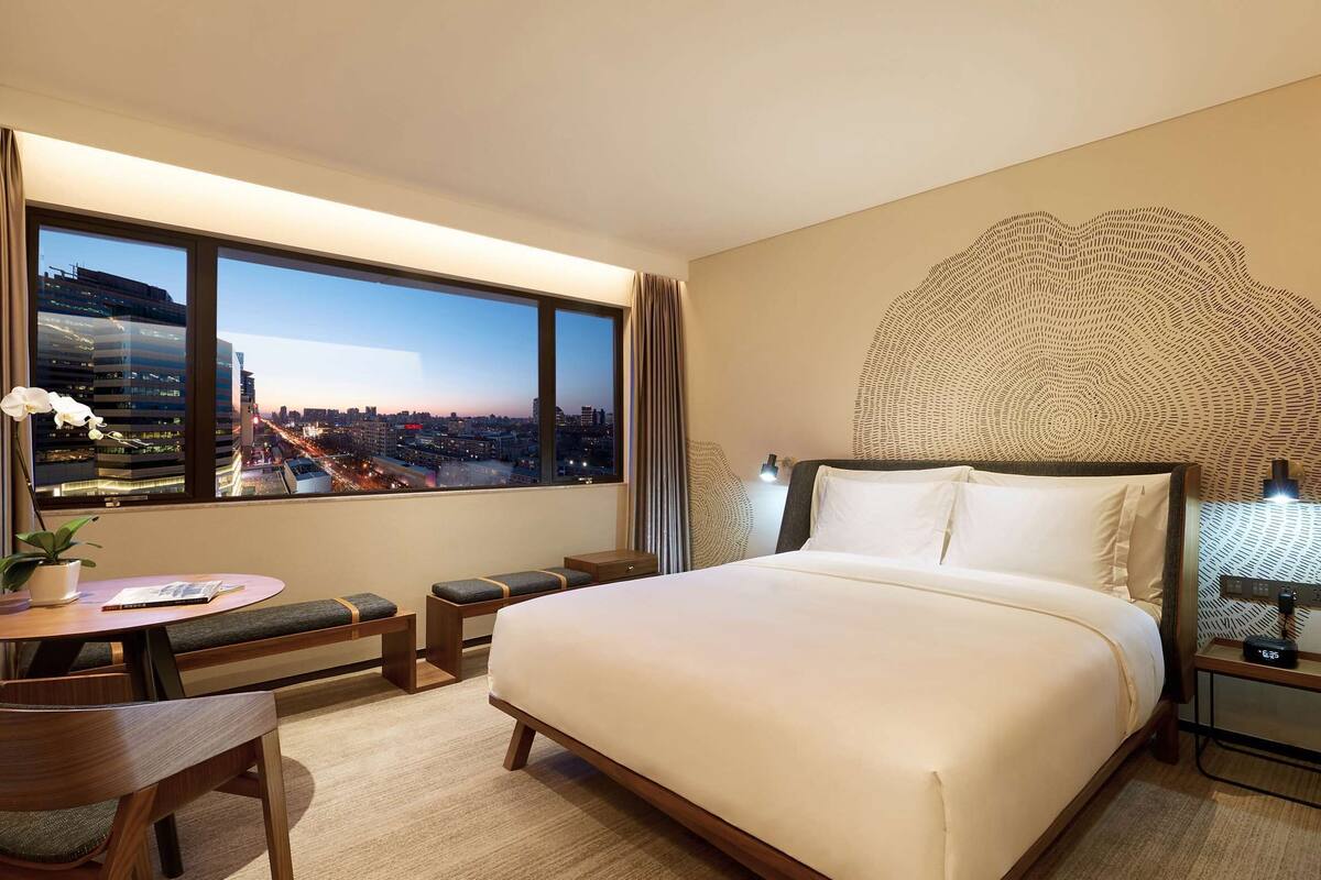 A hotel room with a large bed, a desk with a chair, a small table with a plant, a nightstand with a lamp, and a view of the city at dusk.