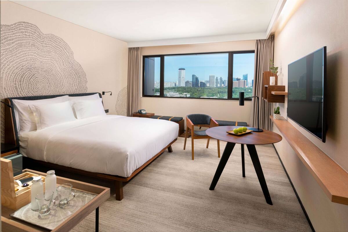 A hotel room with a large bed, a desk with a chair, a TV, and a window with a city view.