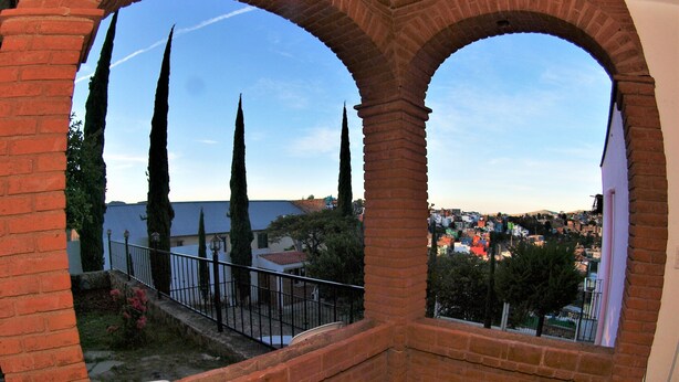 Vista a través de un arco de ladrillo que da a un pueblo con altos cipreses y cielo despejado.