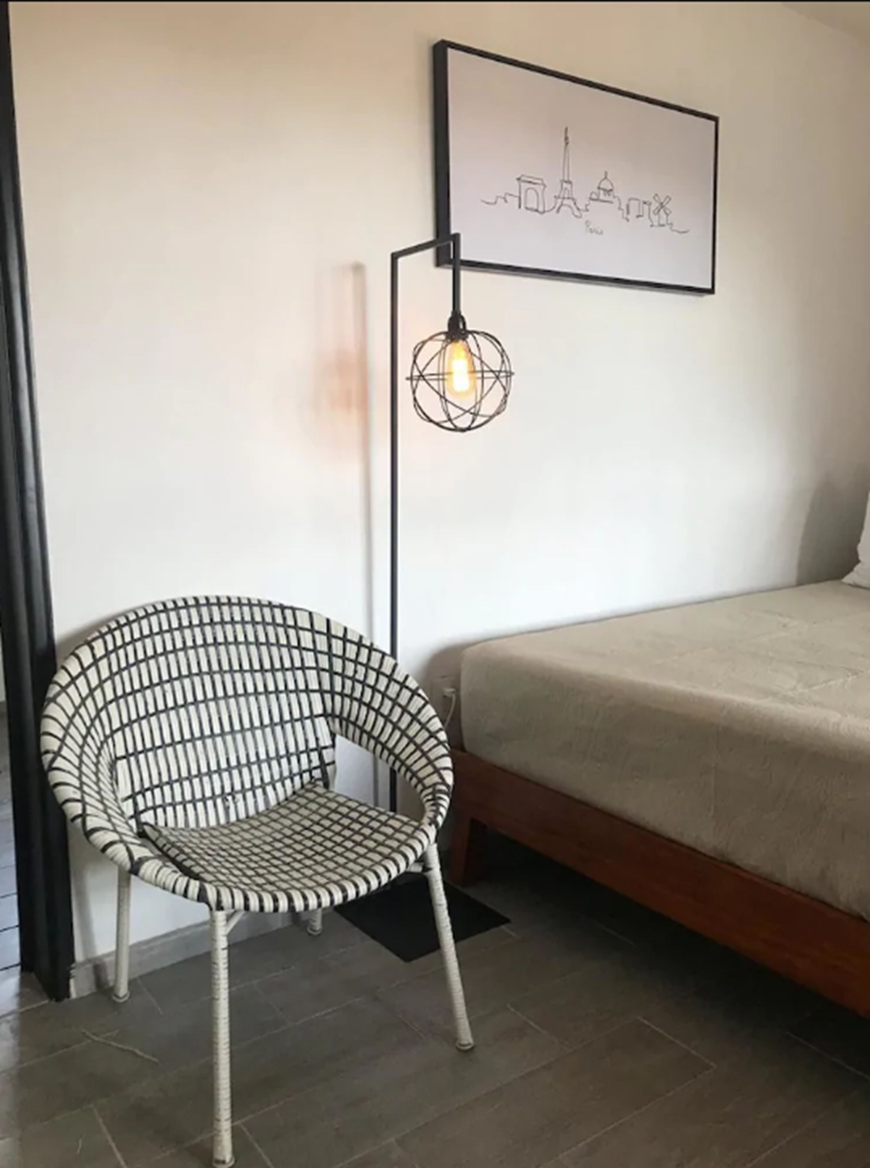 Una habitación con una cama, una silla de mimbre, una lámpara de pie y un cuadro en la pared.