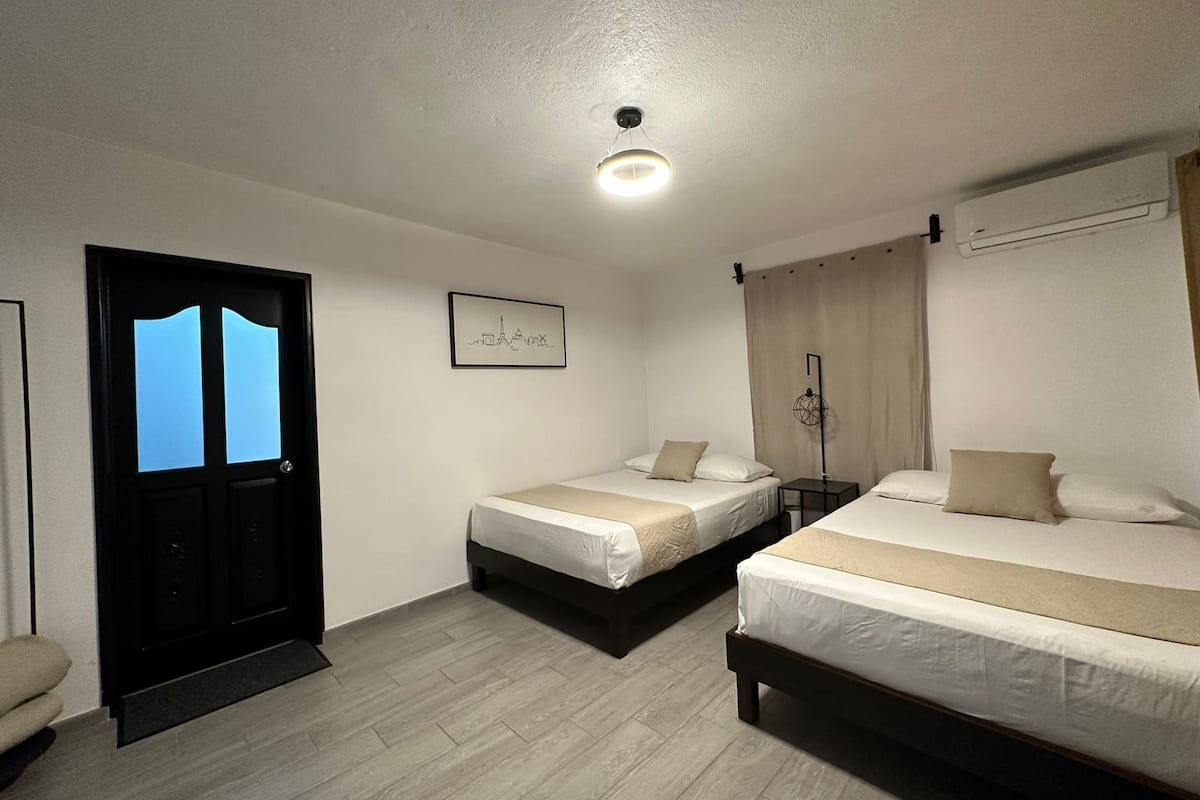 A hotel room with two single beds, a small table, a bedside lamp, and a door with a lighted window.
