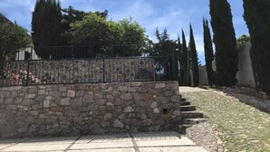 Un muro de piedra con una valla metálica, escalones que suben a un nivel superior y altos cipreses al fondo.
