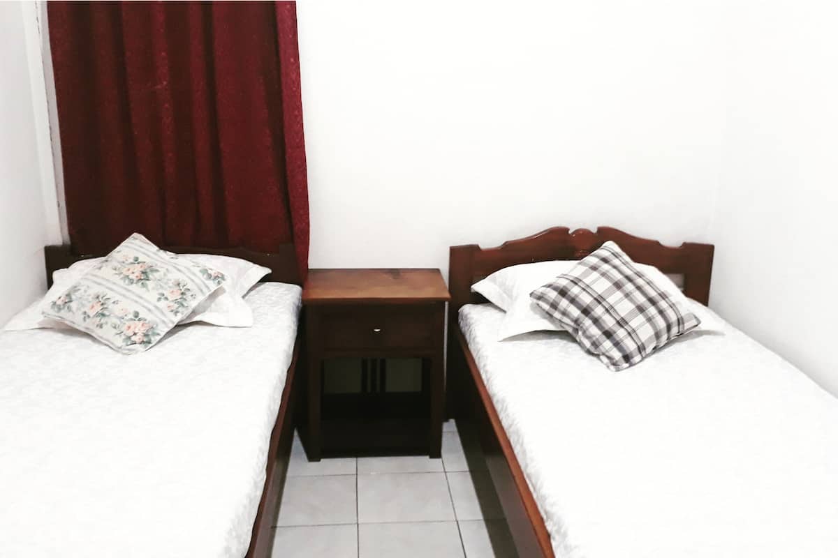 A small, single-room bedroom with two single beds, each with a pillow and a bedside table.