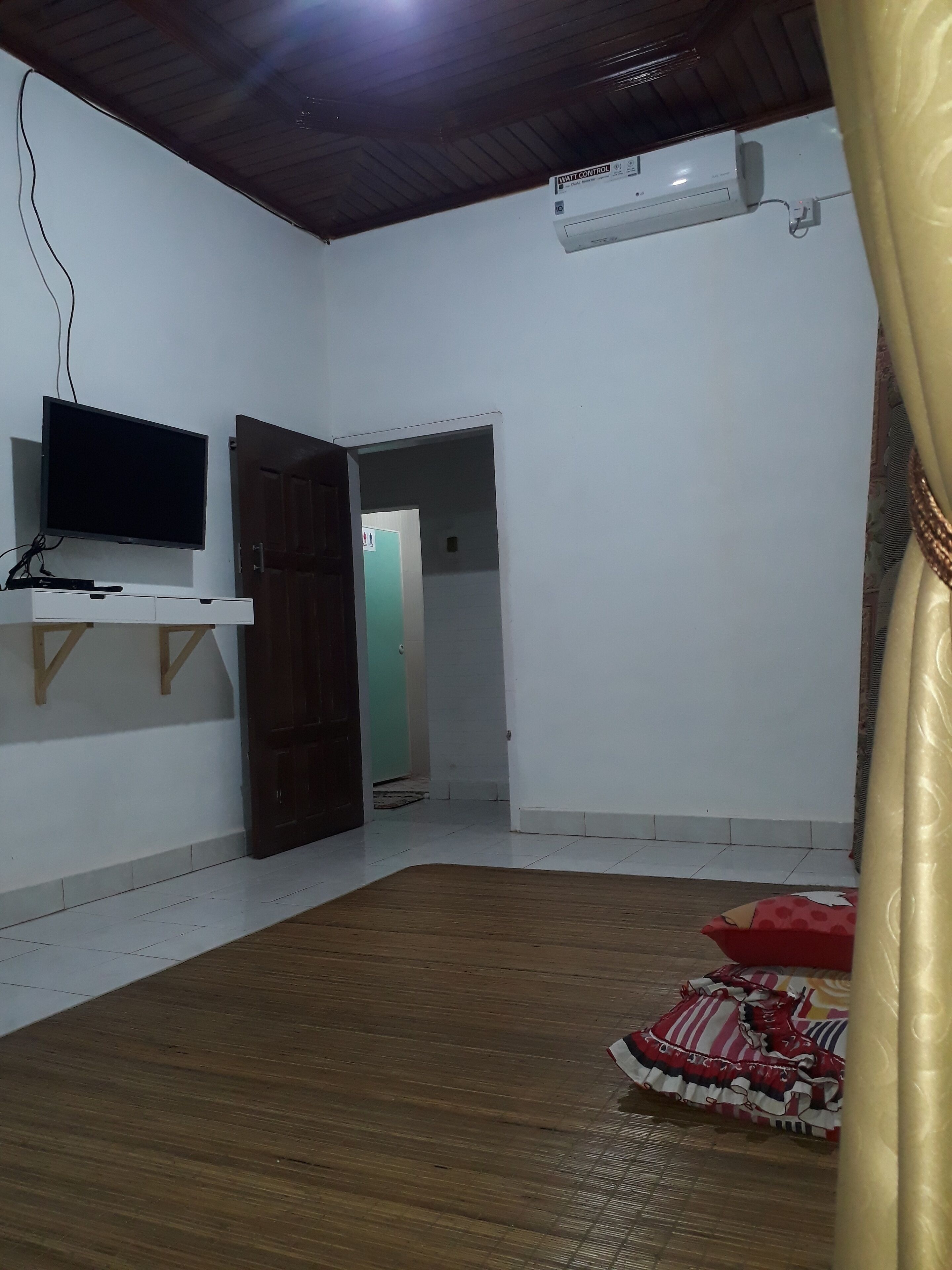 Una habitación con una televisión de pantalla plana sobre un estante blanco, piso de madera y una alfombra con diseño rojo y blanco.