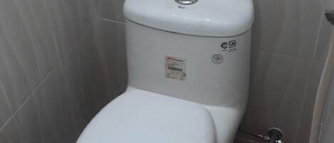 Et lille, kompakt badeværelse med et hvidt toilet, en synlig vandtank og et flisegulv.