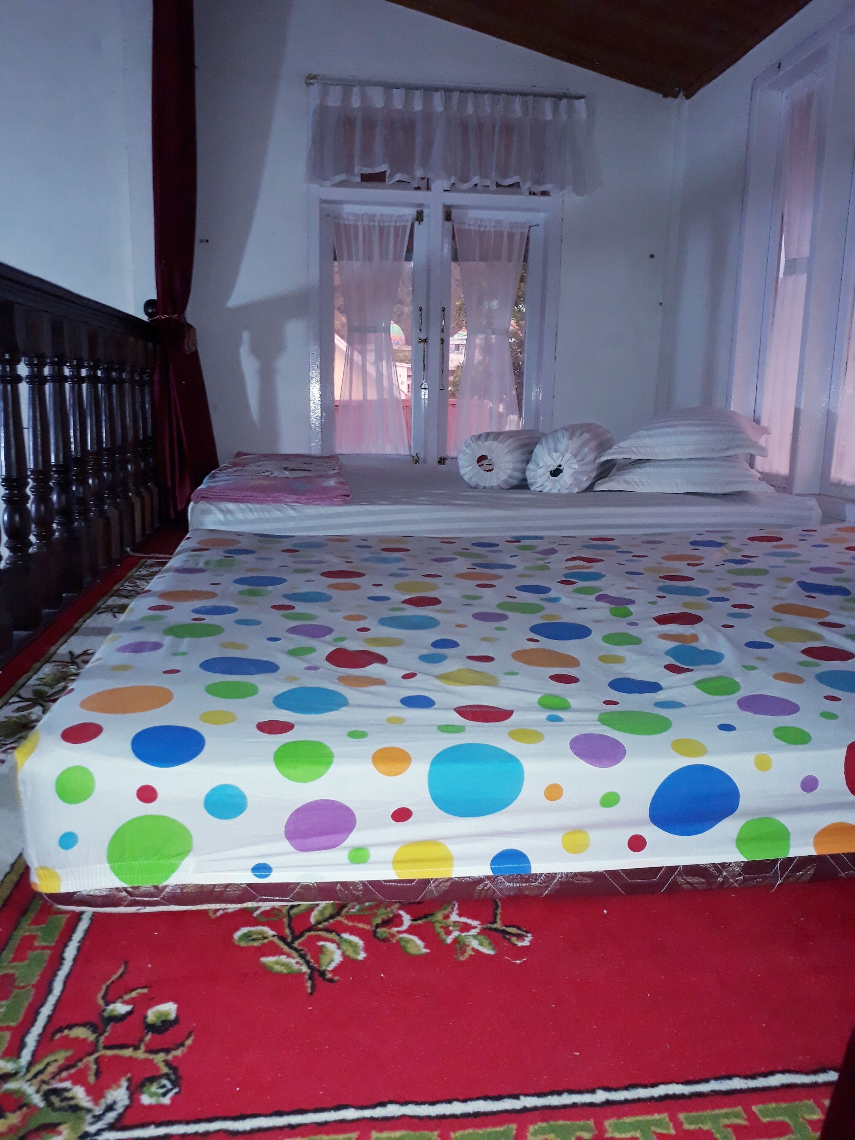 Een slaapkamer met een bed voorzien van een stippenbedekking, een rood tapijt en een raam met dunne gordijnen.