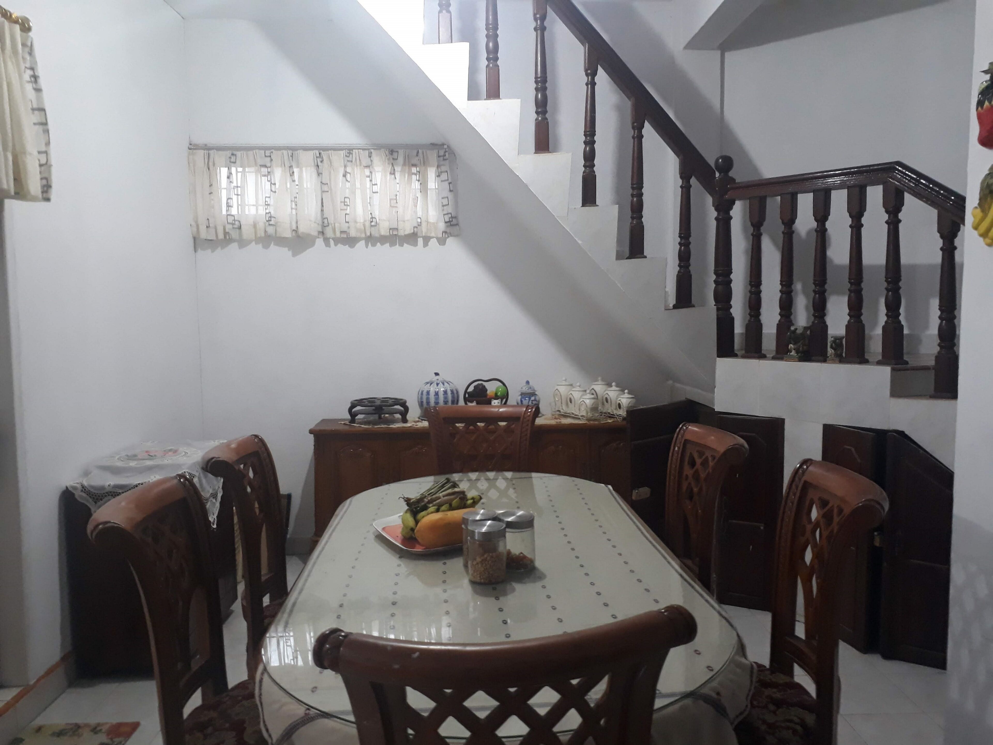 Uma sala de jantar com uma mesa de vidro, cadeiras de madeira, uma escada com balaústres de madeira e uma janela com cortinas estampadas.