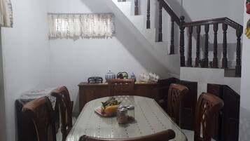 Uma sala de jantar com uma mesa de vidro, cadeiras de madeira, uma escada com balaústres de madeira e uma janela com cortinas estampadas.
