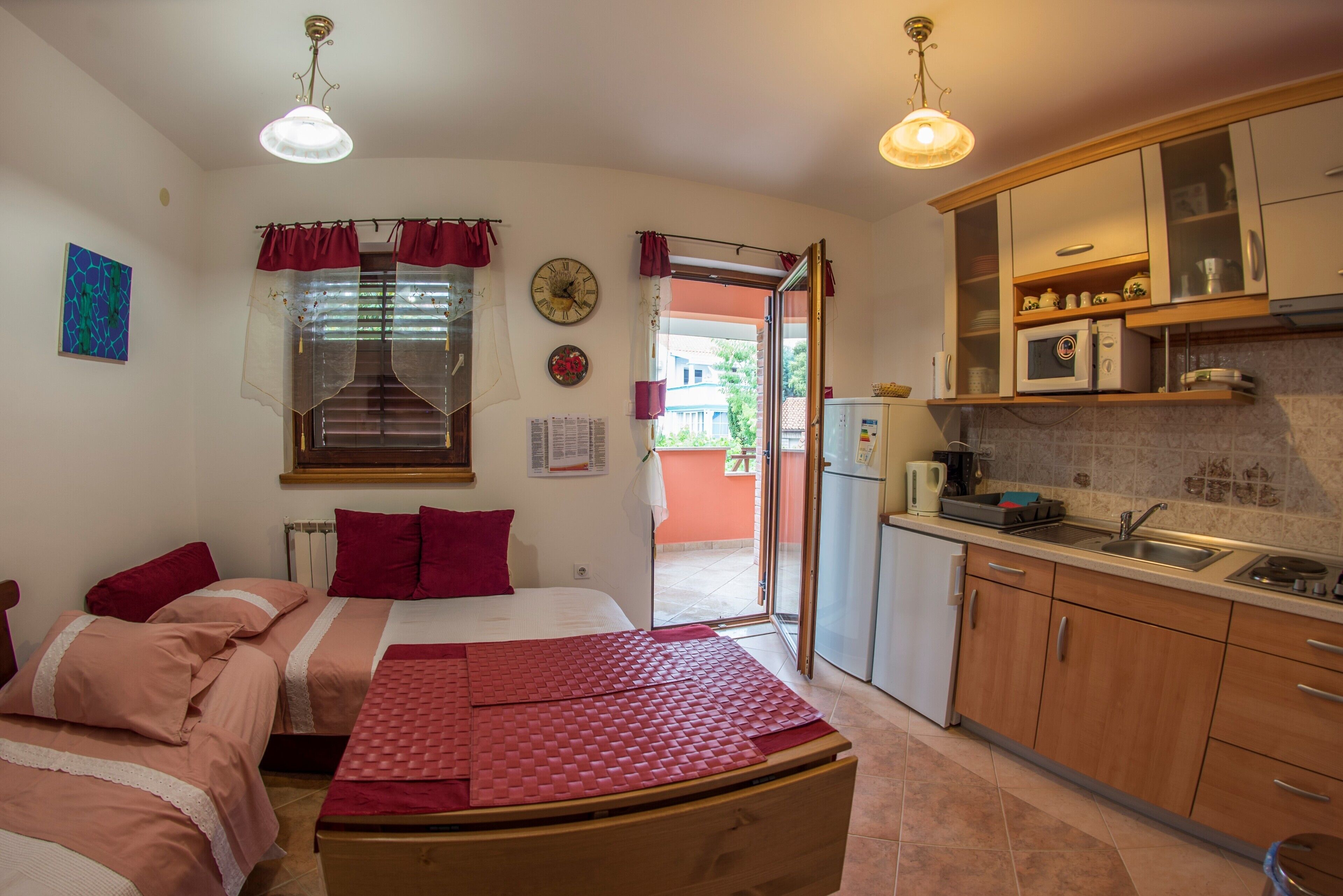 Een slaapkamer met twee bedden, een houten bankje en een rode deken. Er is een keuken met houten kastjes en een magnetron.
