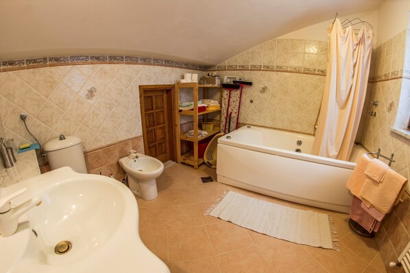 Un baño con bañera, bidé, lavamanos, un estante de madera con artículos de aseo y cortina de ducha.