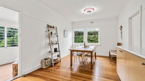 Ein Zimmer mit Holzboden, weißen Wänden, einem Esstisch mit Stühlen, einem Fenster mit Blick ins Grüne und einer Deckenleuchte.