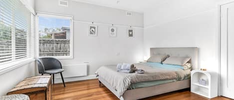 Une chambre à coucher avec un lit, une chaise, une commode en bois et une fenêtre avec des stores.