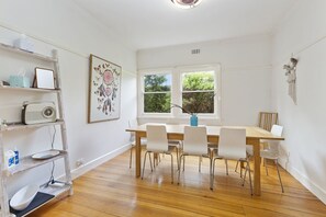 A dining area with a wooden table, white chairs, a shelf with items, and a window with a view of greenery.