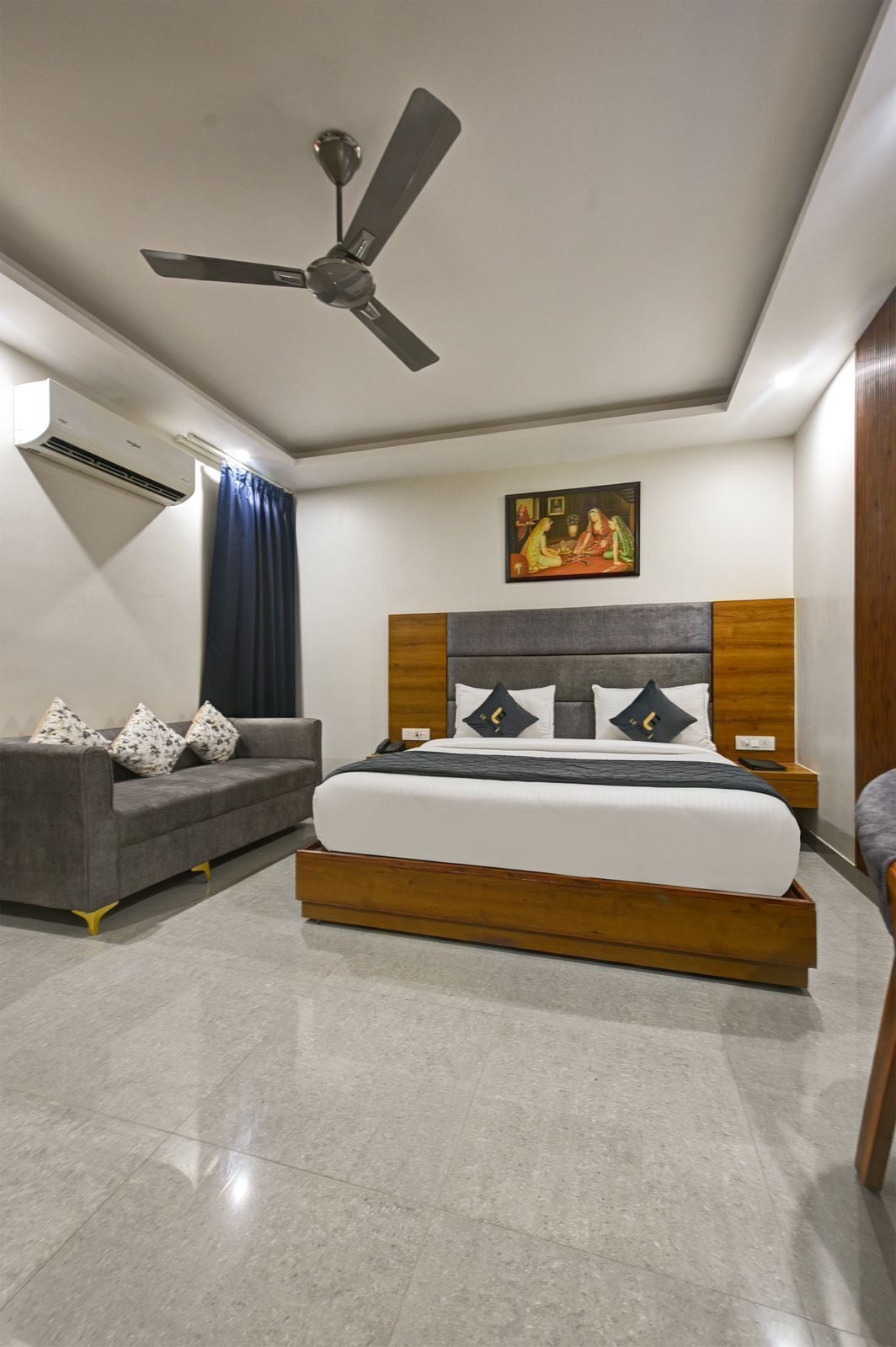 A bedroom with a bed, a grey sofa, a ceiling fan, an air conditioner, and a painting on the wall.
