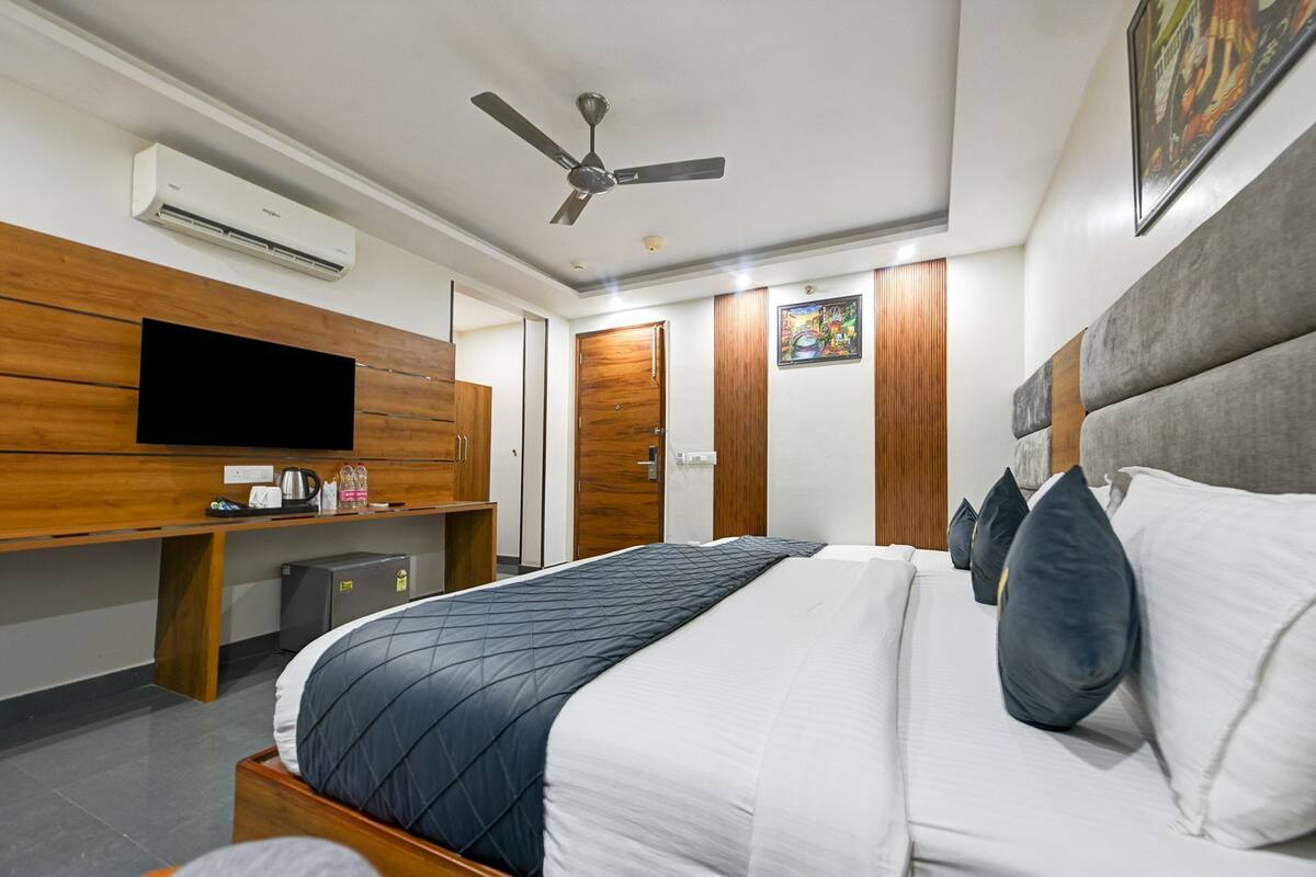 Quarto de hotel com cama, televisão, ventoinha no teto e uma pequena escrivaninha com chaleira e chávenas.