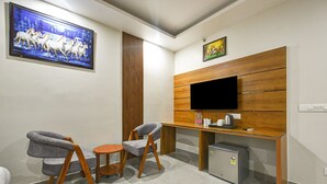 A modern room with a flat-screen TV mounted on a wooden panel wall, two grey upholstered chairs, a small round table, a mini fridge, and a painting of horses on the wall. -  Hotel Smart by Kelvish Hotel (New Delhi)