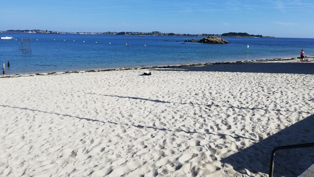 A sandy beach with clear blue water, a rocky outcrop, and a distant shoreline with buildings. - Studio with garden at the doors of the beach and thalasso ROSCOFF (Roscoff)