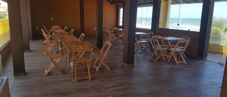An indoor dining area with wooden tables and chairs, large windows with a view of the ocean, and a tiled floor.